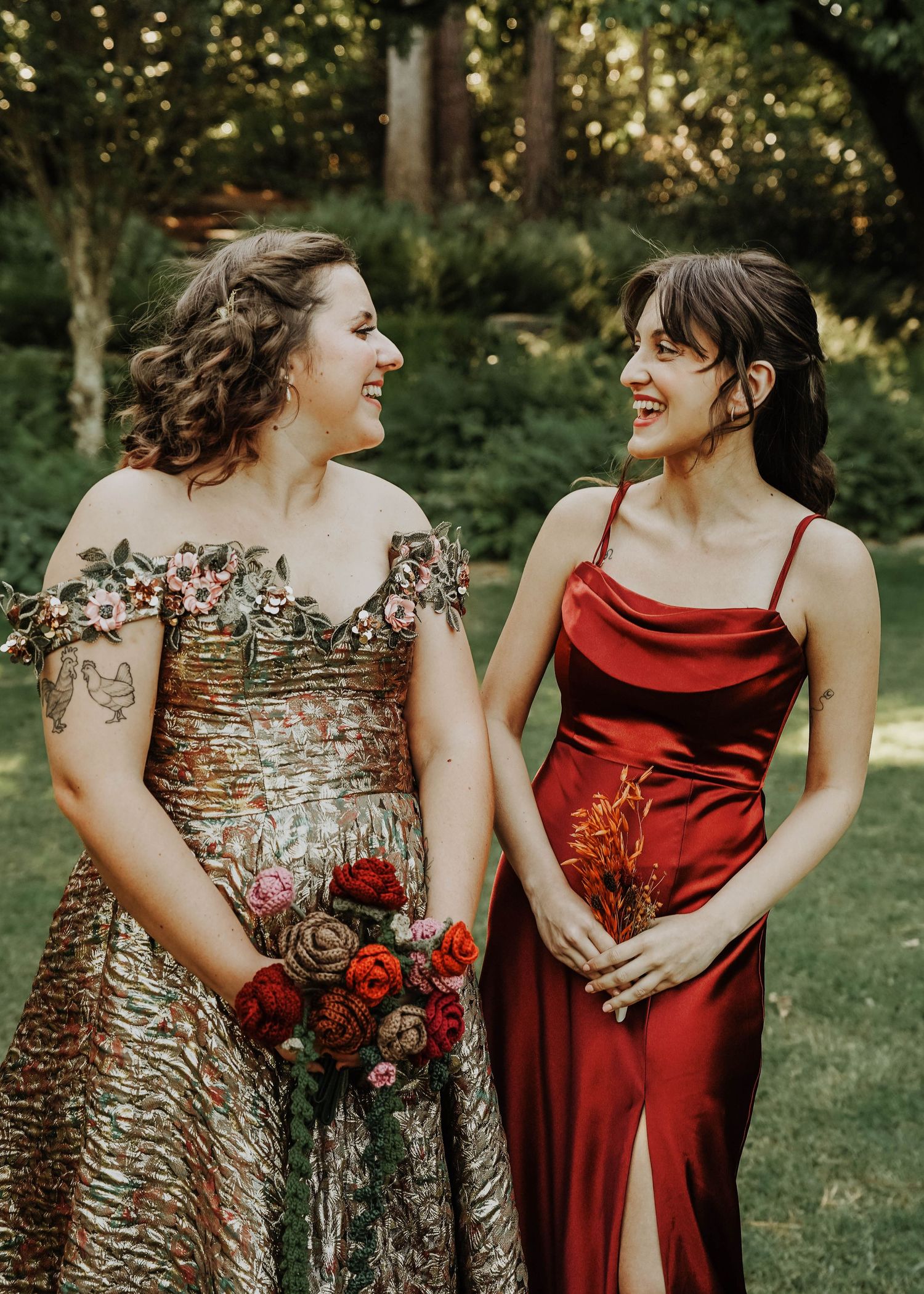Two friends in formal dresses share a laugh while holding red roses in garden setting.
