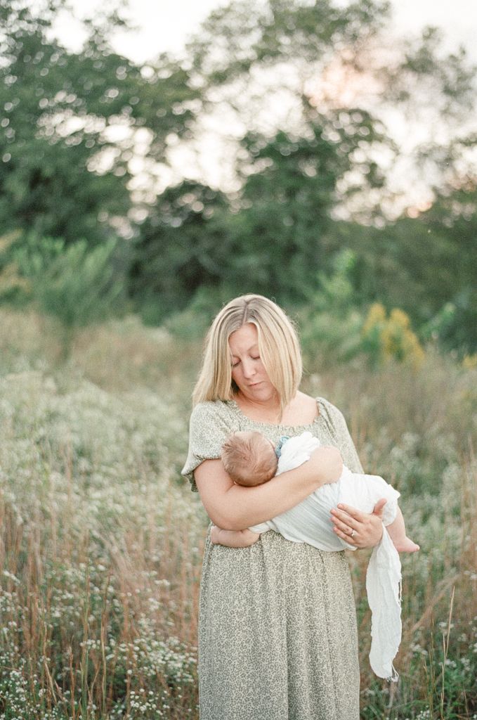 Mother smiles down at her baby as they stand in a field at sunset