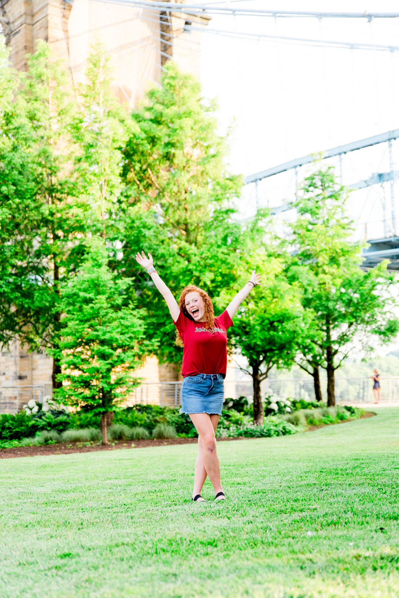 young woman in Transylvania shirt and jean skirt putting arms int he air in front of Roebling Bridge at Smale Park