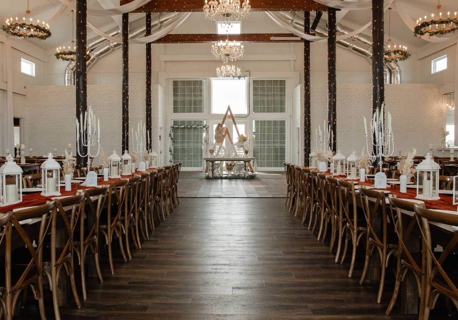 Elegant wedding reception venue with long wooden tables, chandeliers and white decor in rustic style.