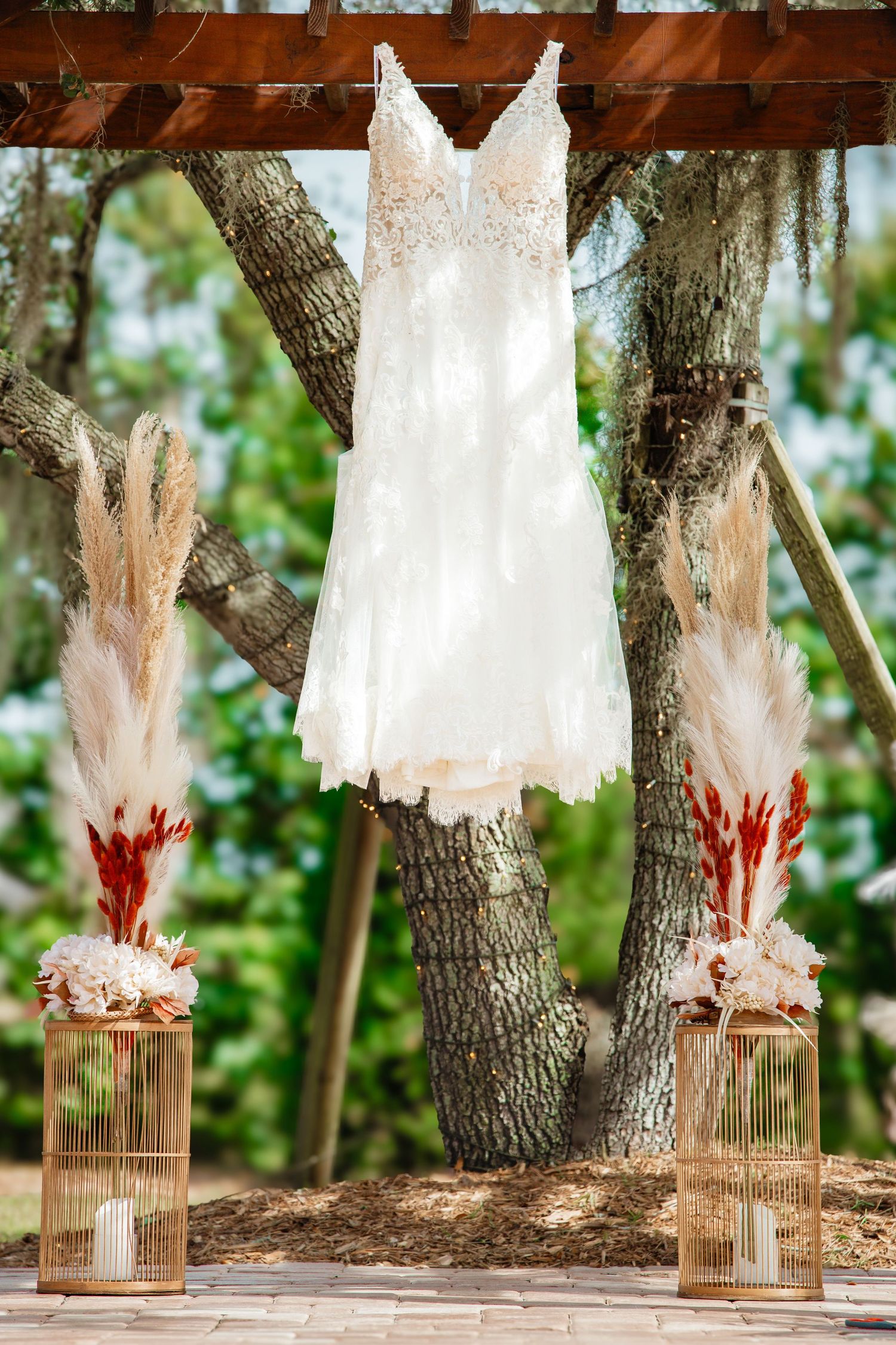 A white lace wedding dress hanging from a tree with decorative pink and red floral arrangements in glass vases below.