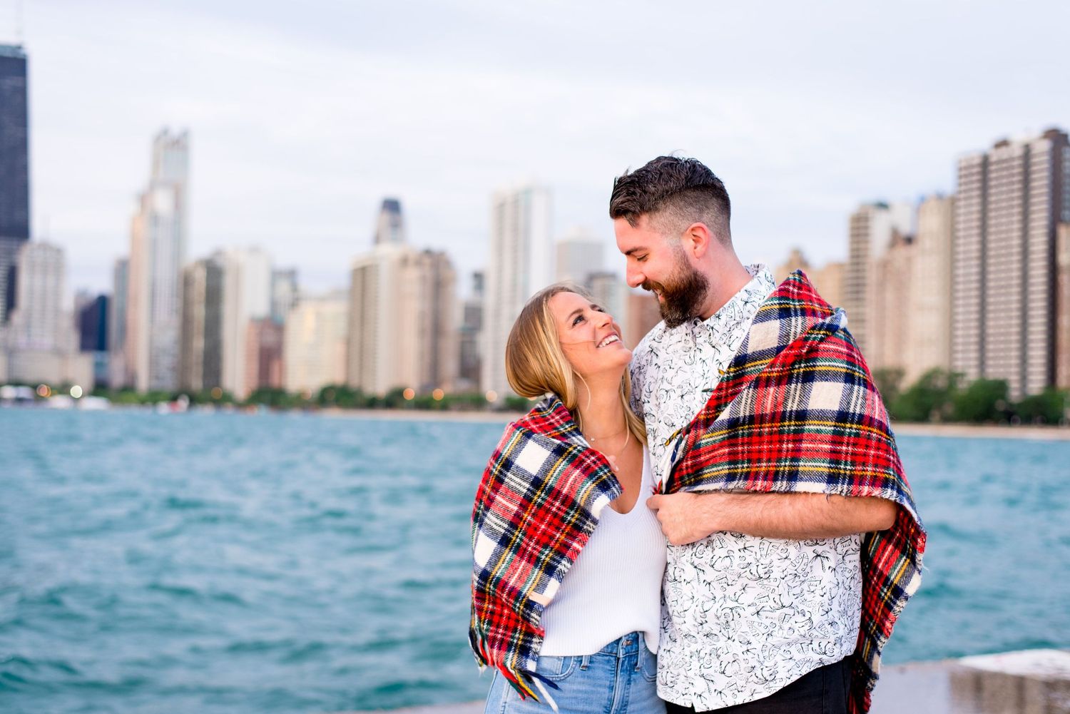 young engaged couple smiles at each other and wraps up in a red plaid blanket by the water at North Avenue Beach