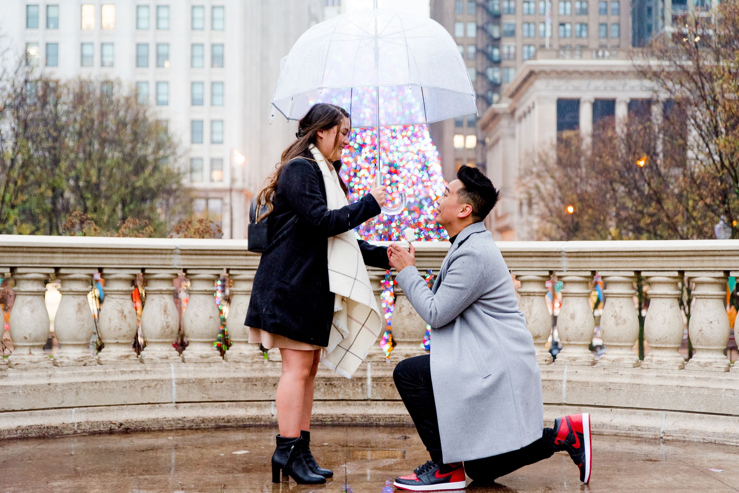 man in grey coat kneeling proposing to woman in black coat and clear umbrella in front of Millennium Park Christmas Tree