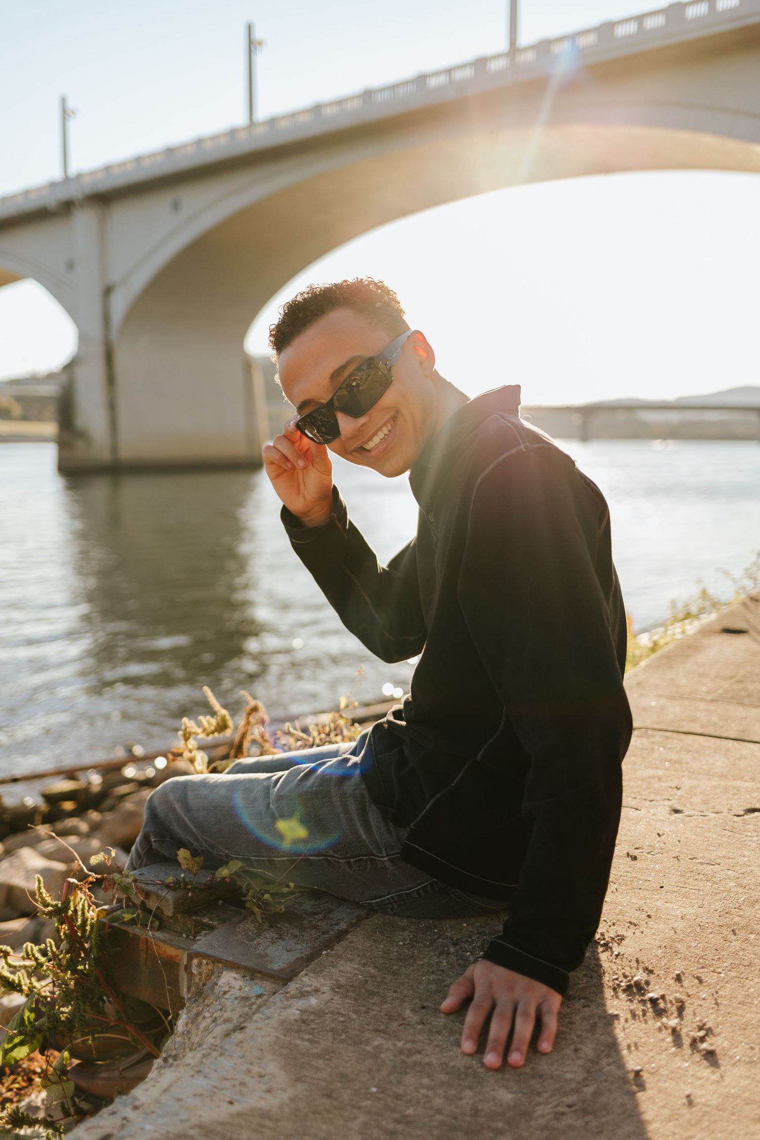 A young man sits next to a river and tips his sunglasses towards the camera