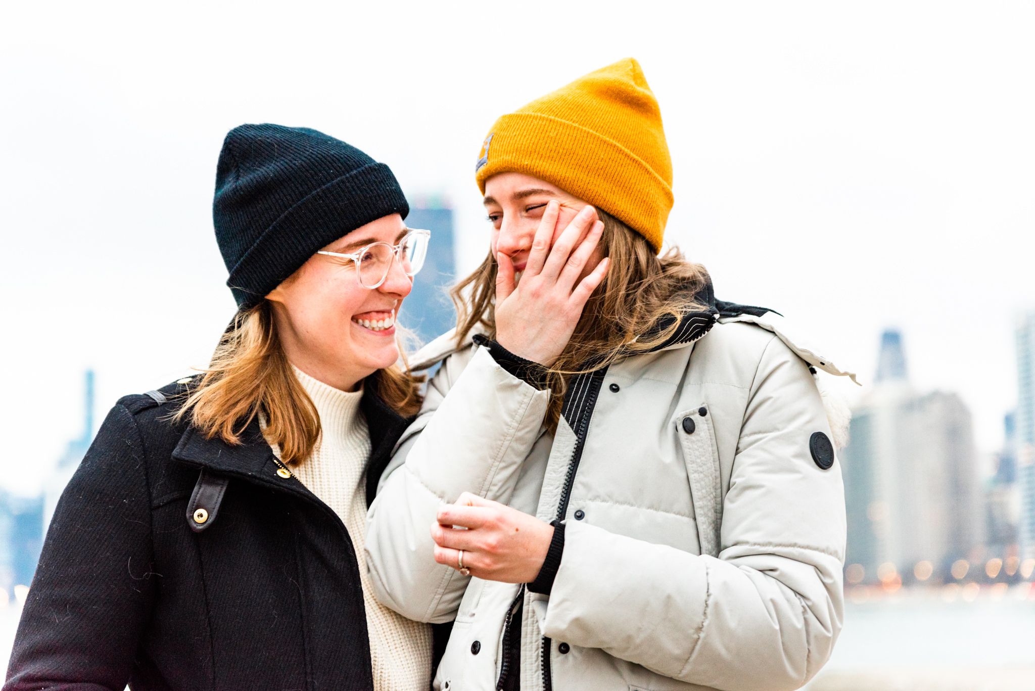 woman in yellow hat crying and fiancee in black hat and coat smiling at her after Chicago proposal at North Avenue Beach
