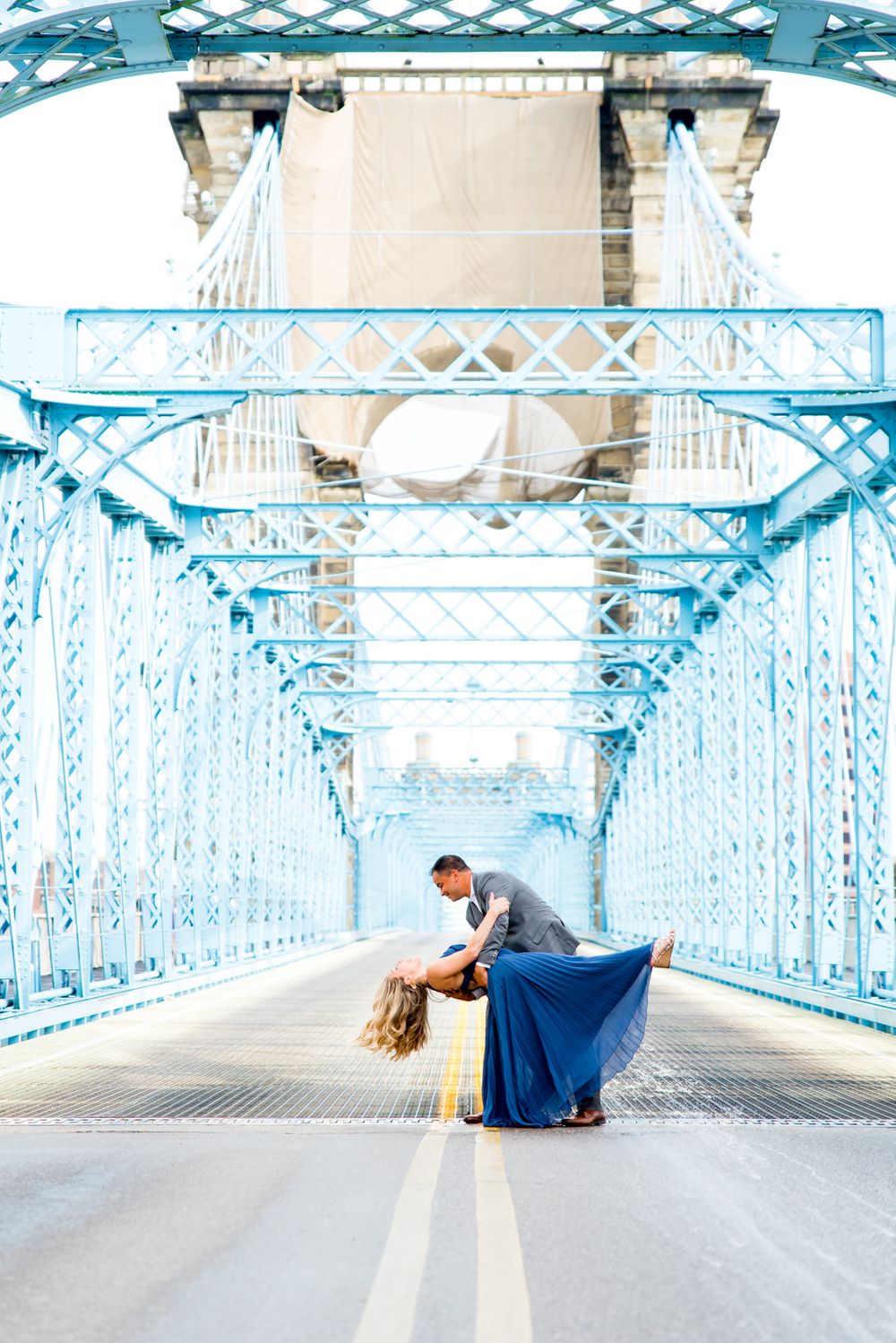 man in gray suit dips woman in blue dress in the middle of Roebling Bridge for Cincinnati engagement pictures