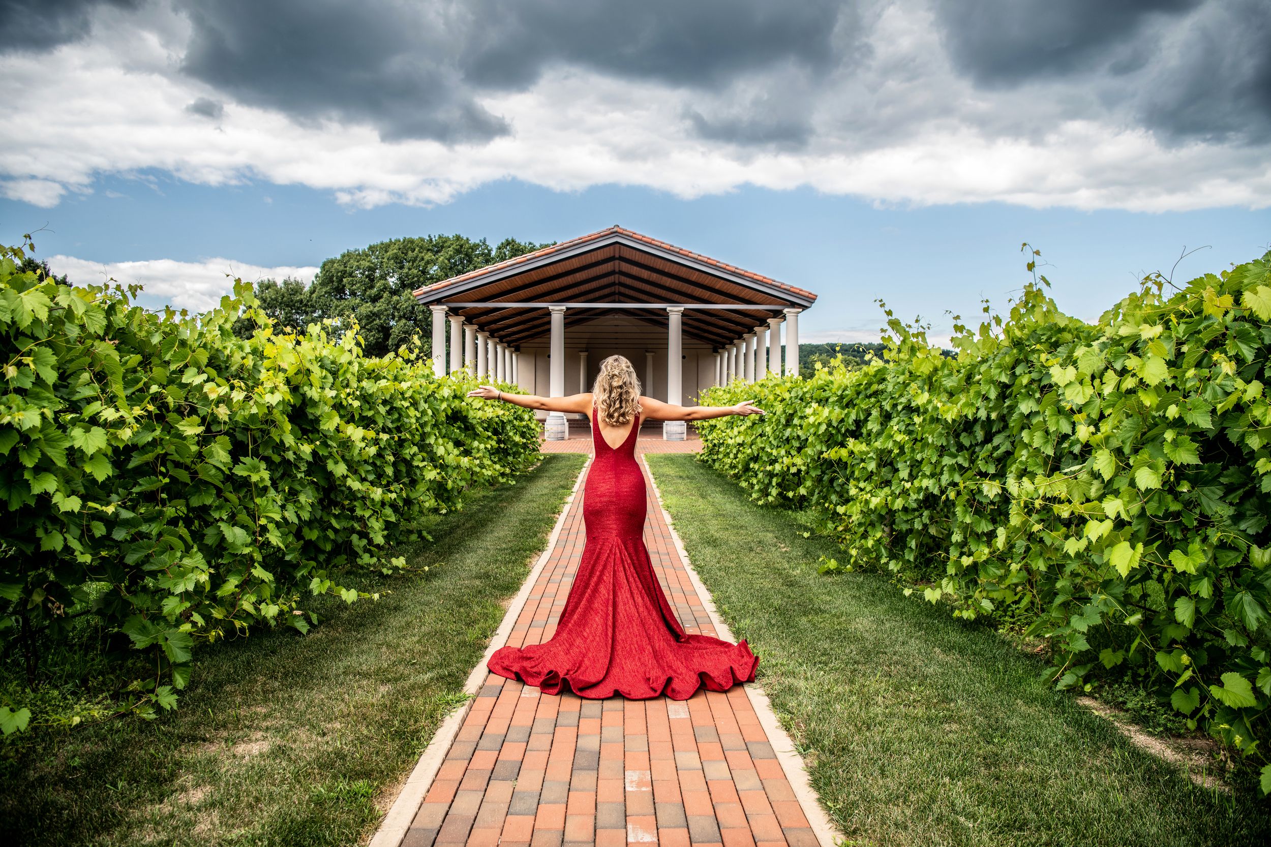 Senior Girl posing in red prom dress in vineyard, arms outstretched