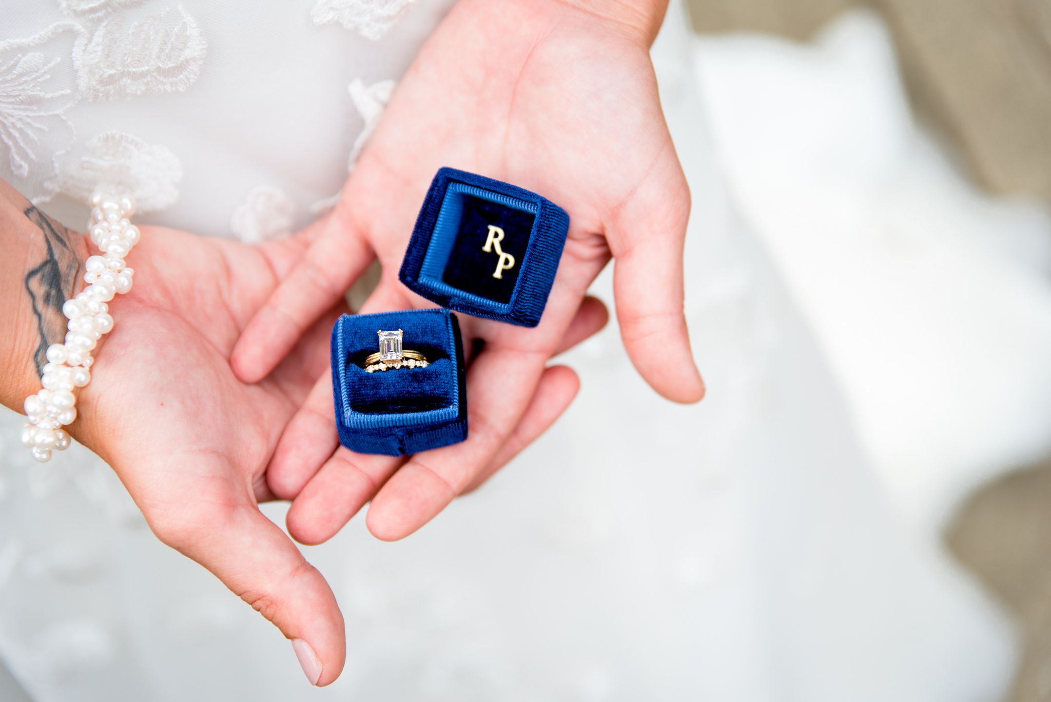 bride with wrist tattoo and pearl bracelet holding blue ring box with emerald cut diamond ring inside
