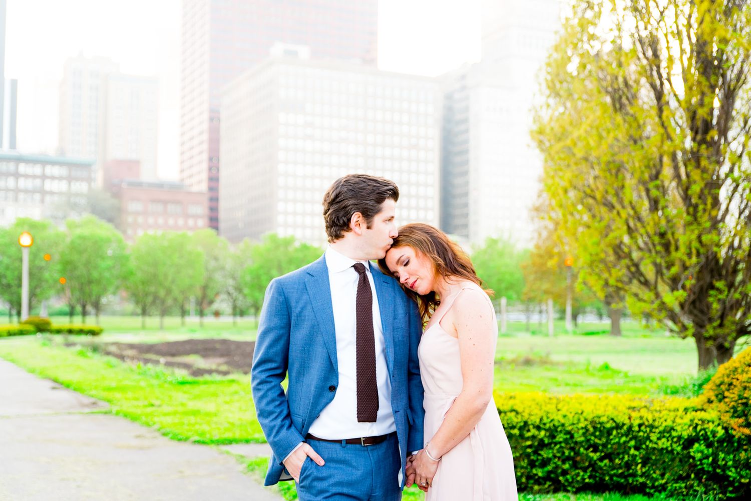 man in Navy suit kissing fiancee in pink dress on forehead in Chicago Grant Park for sunset engagement pictures