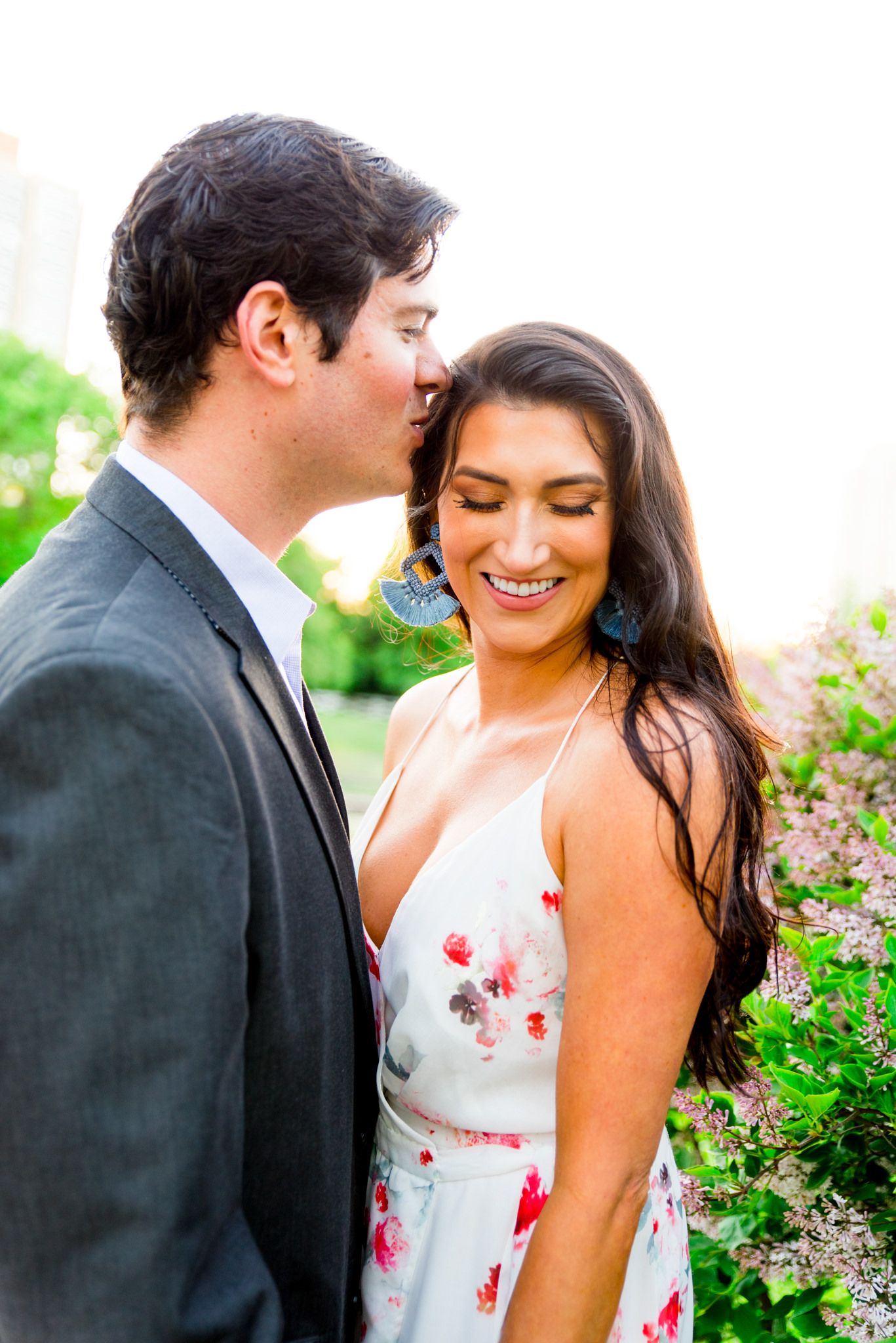 man in gray suit kissing woman in floral dress and blue earrings at sunset while she smiles for engagement pictures