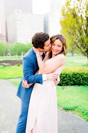 man in navy suit hugs and kisses fiancee in pink dress who is smiling in Millennium Park Chicago engagement