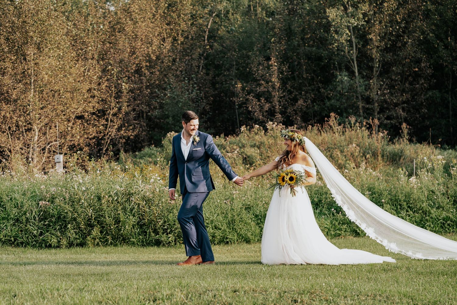 Newlyweds holding hands while walking through sunny meadow with flowing bridal veil.