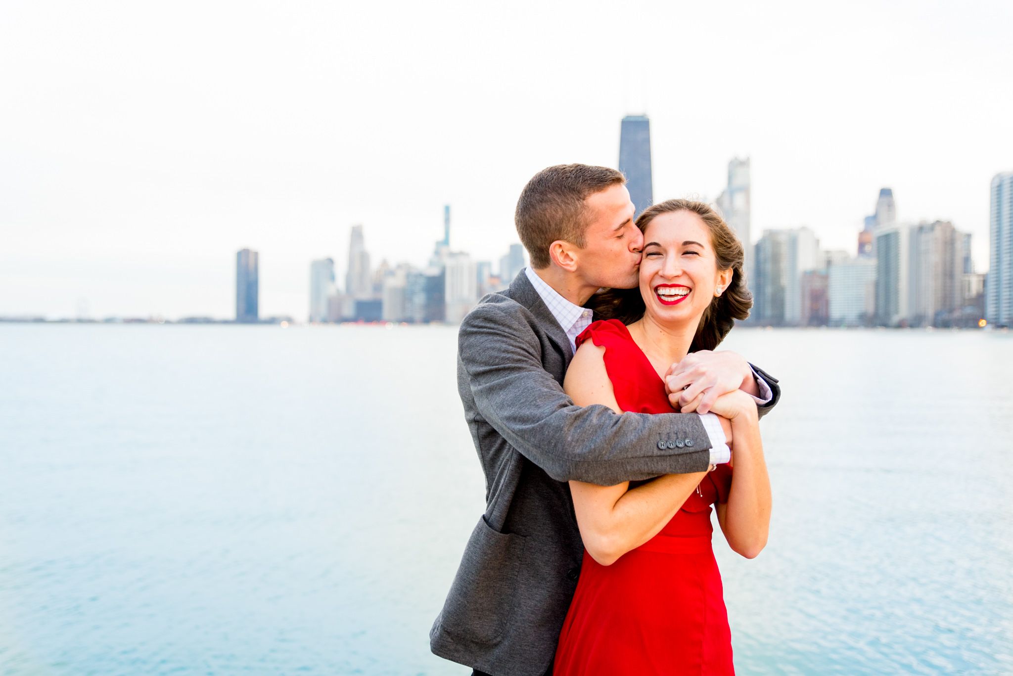 man hugging fiancee from behind and kissing her cheek in front of Chicago skyline for Chicago engagement pictures