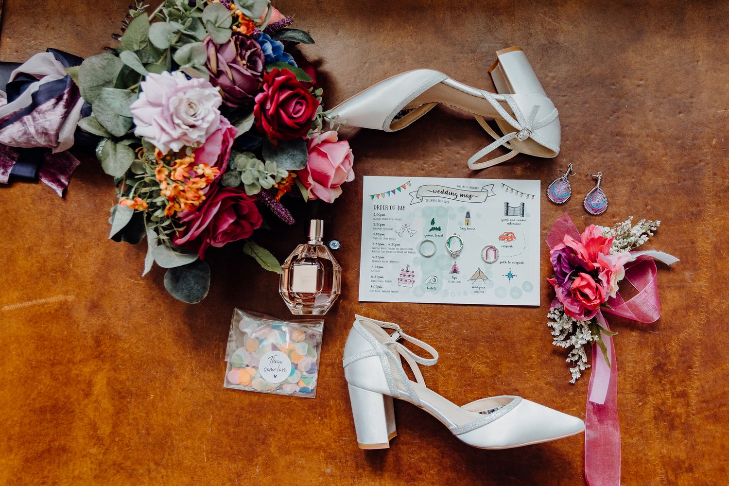Wedding details flatlay with white heels, floral bouquet and invitation on wooden surface.