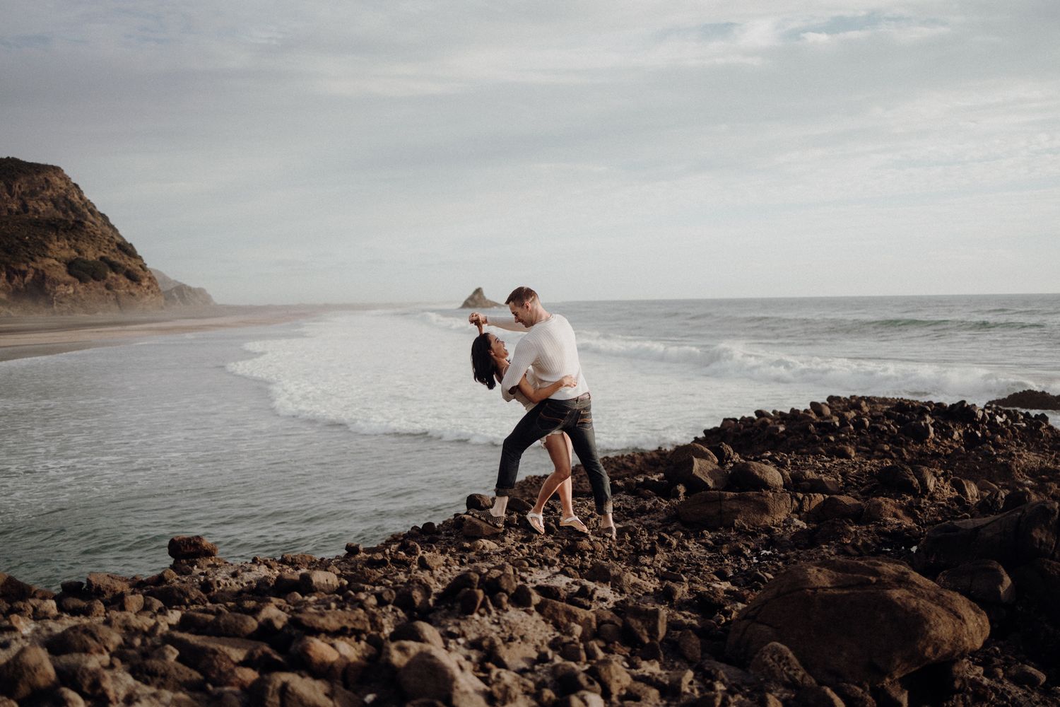 A couple explores rocky coastal terrain while holding hands near ocean waves.