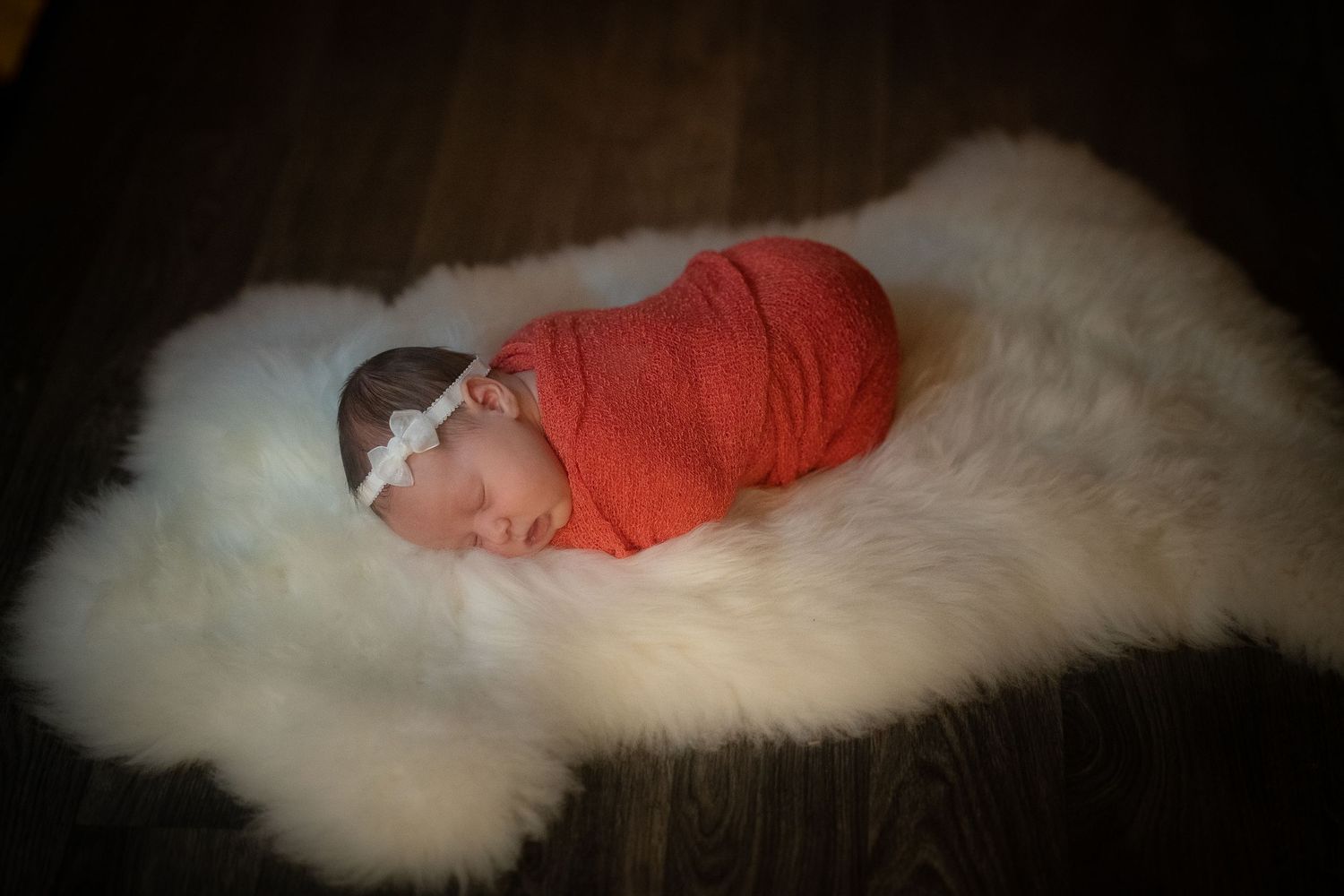 A newborn baby swaddled in coral fabric sleeping peacefully on white fur.