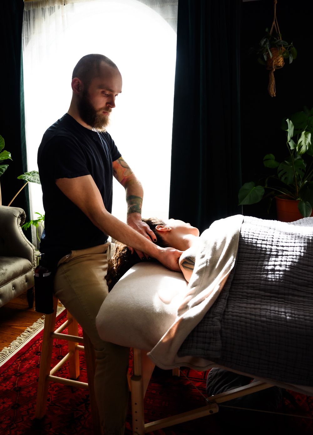 A massage therapist sitting on a stool while stretching a person's neck as they lie face up looking relaxed in daytime
