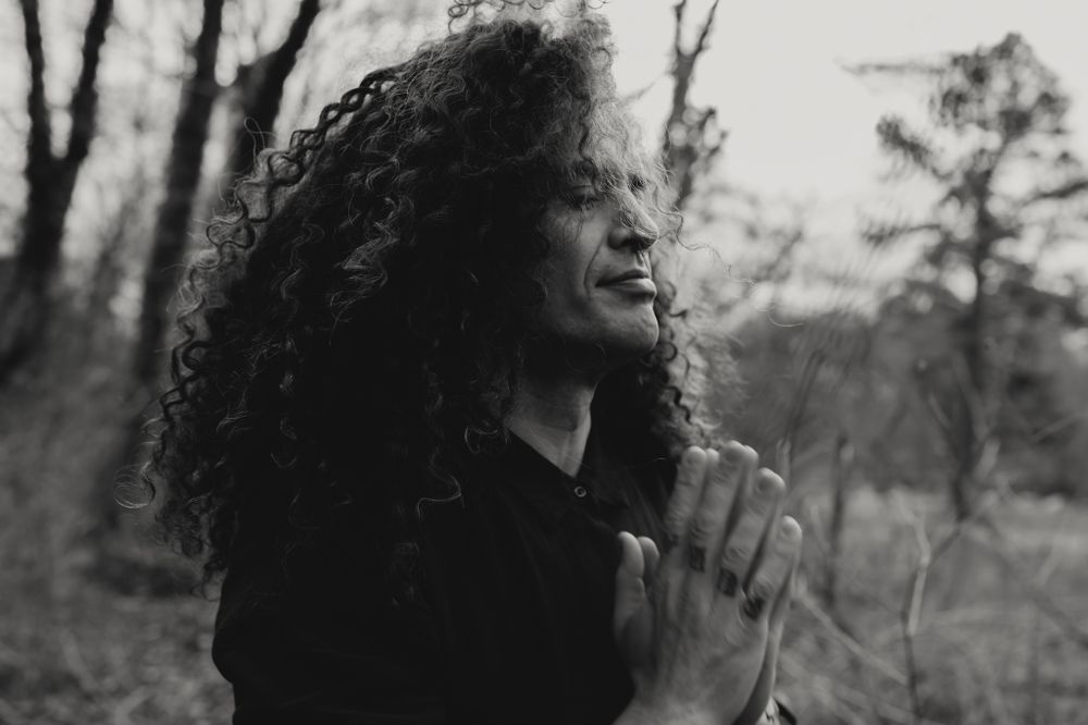 A person with long curly hair outside with their eyes closed and hands pressed together palms facing in black and white
