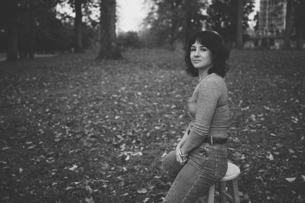 A portrait of a woman in black-and-white as she sits on a wooden stool in a field of fallen leaves smiling coyly