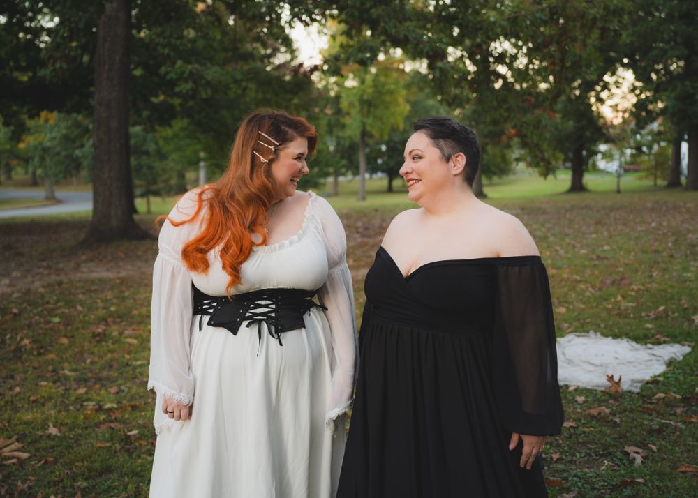 Two friends looking at each other and laughing wearing long formal dresses in a field at dusk