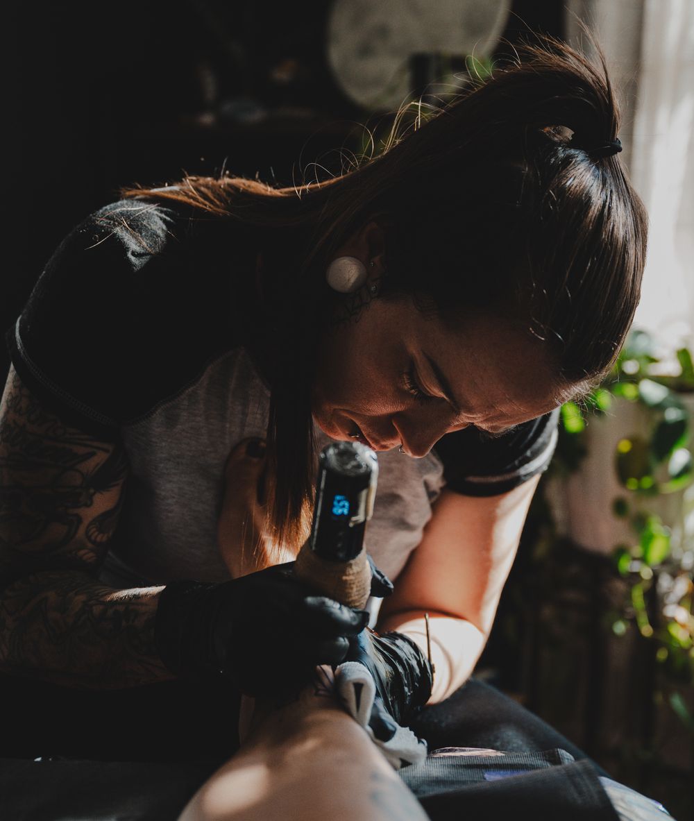 A person giving a tattoo wearing black gloves with warm natural light inside a room with black walls and vibrant plants