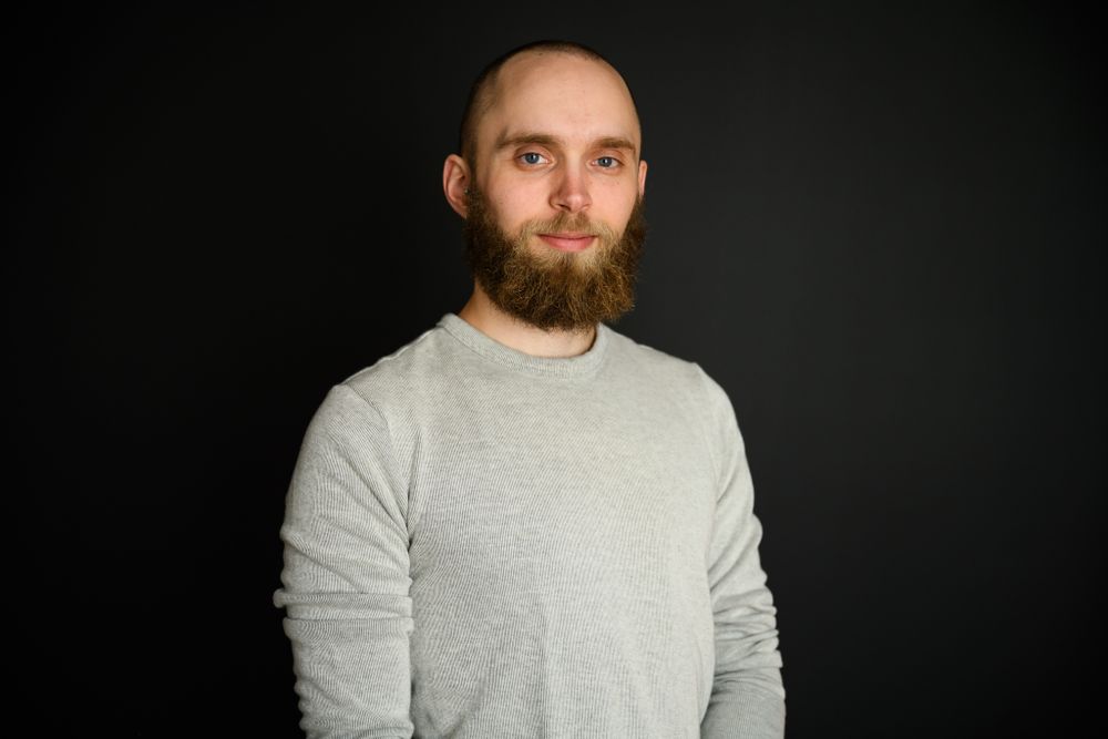 A person with a short haircut and beard in front of a black wall wearing a grey long sleeve shirt and smiling softly