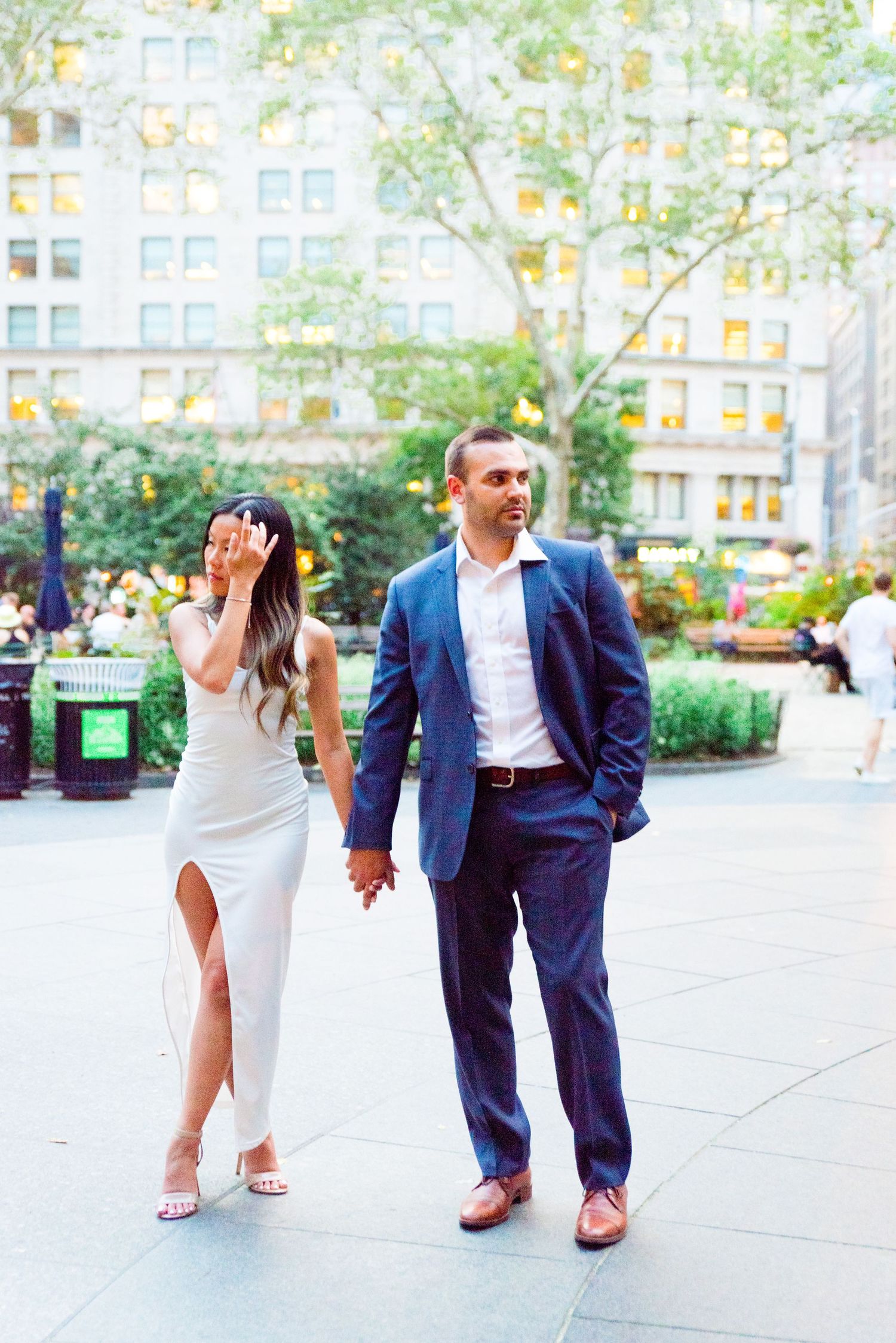 fiances in white and navy hold hands in front of building in Madison Square Park at sunset
