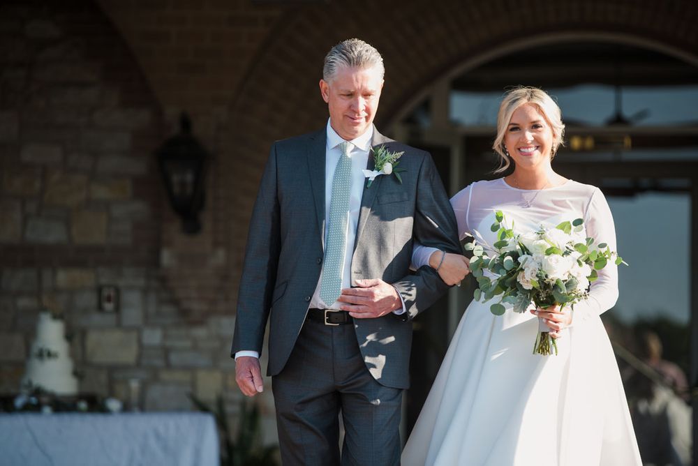 A bride wearing a Robert Bullock Wedding gown is escorted down the aisle by her father wearing a gray suit.