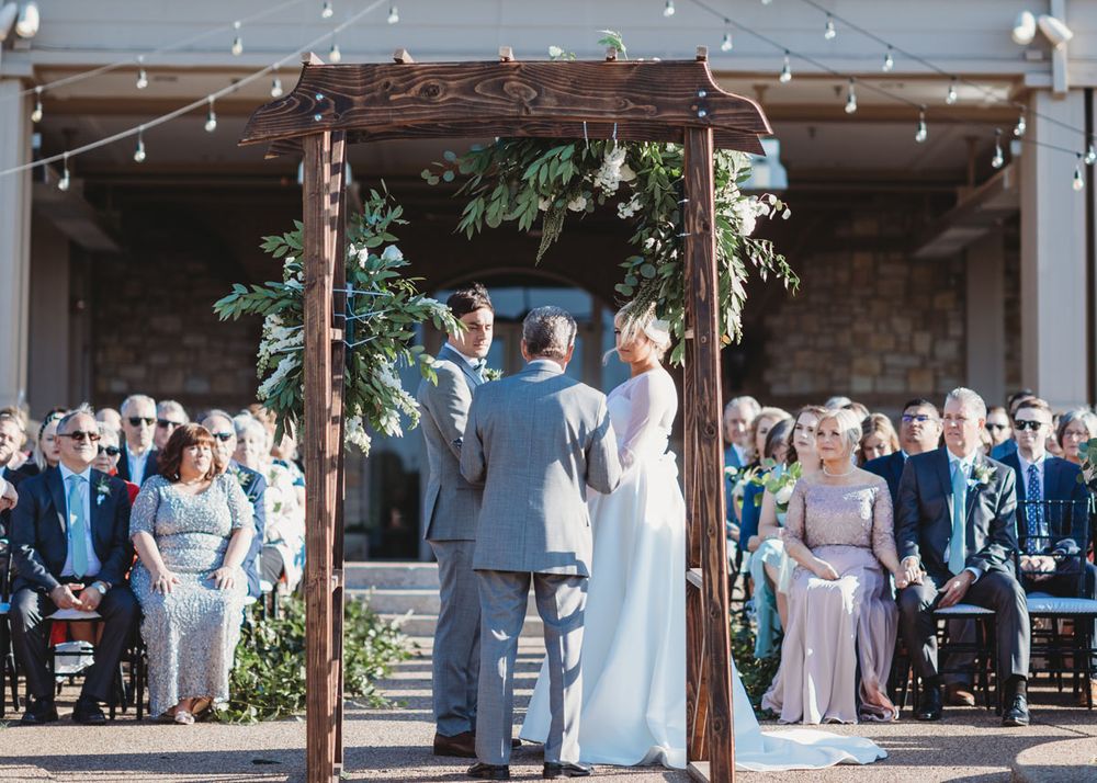 A bride wearing a Robert Bullock wedding gown exchanges wedding vows with groom wearing grey tuxedo at Stone Briar Club