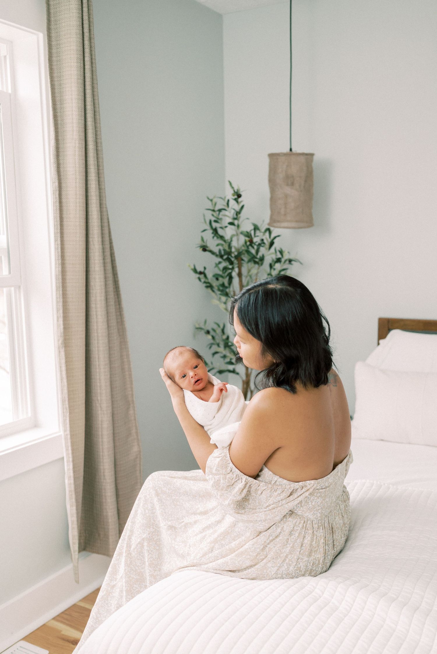 Parent in white dress holding baby near window with natural light streaming in.