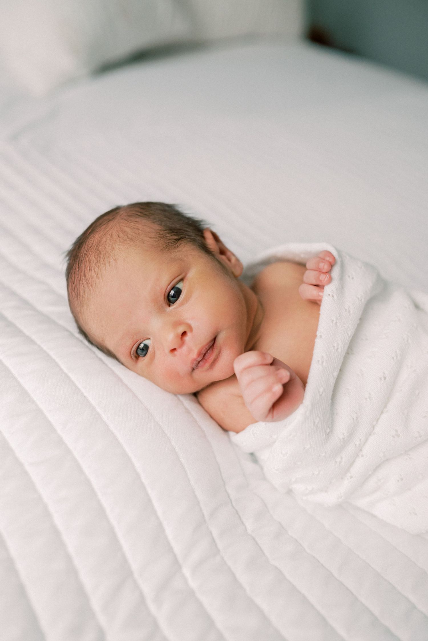 A newborn baby wrapped in white bedding looking at the camera.