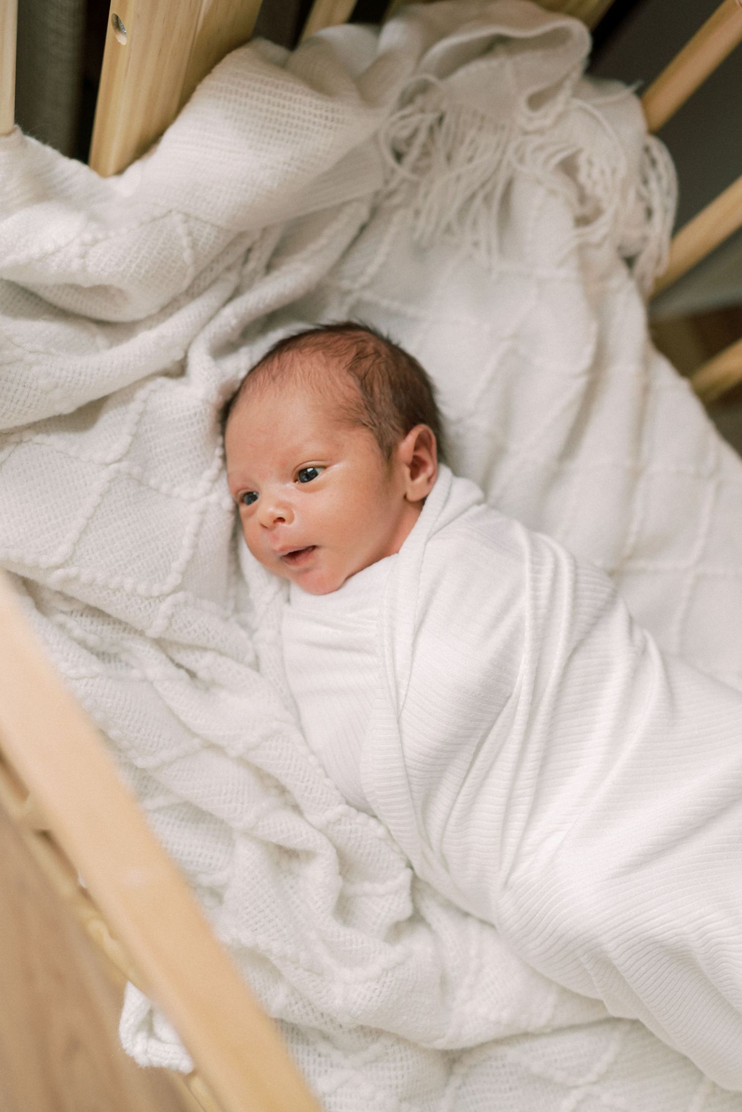 A newborn baby swaddled in white fabric lying on soft textured bedding.
