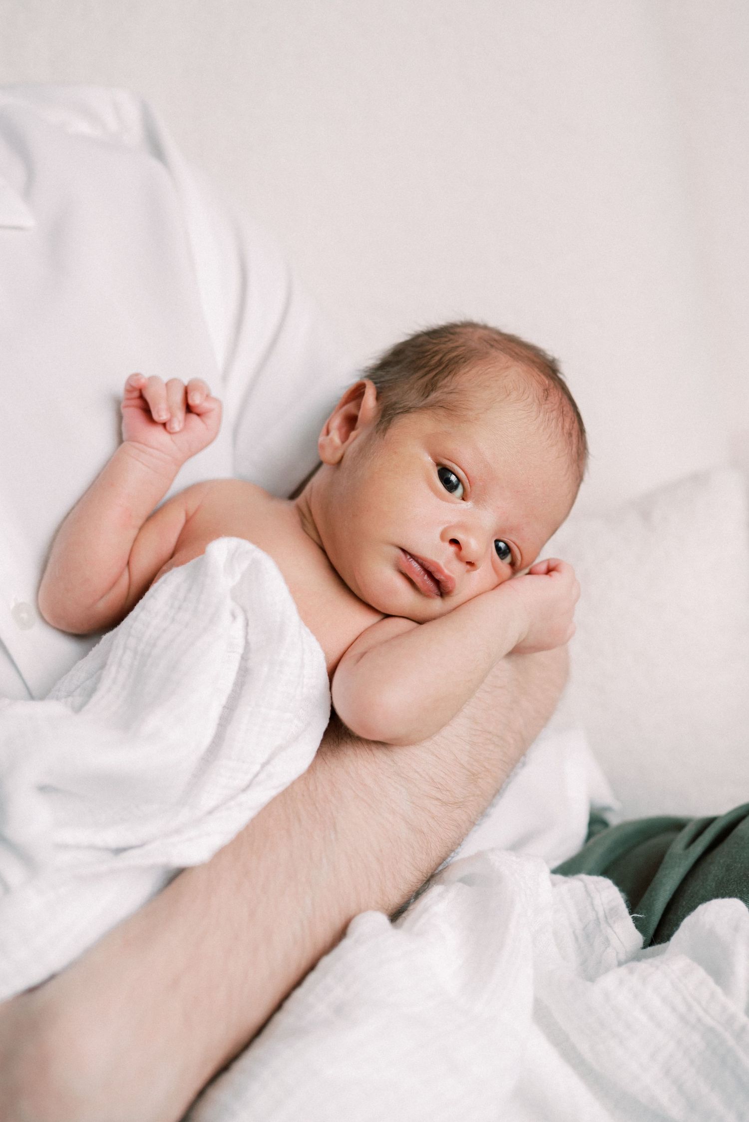 A newborn baby resting on white bedding with bright natural window light.