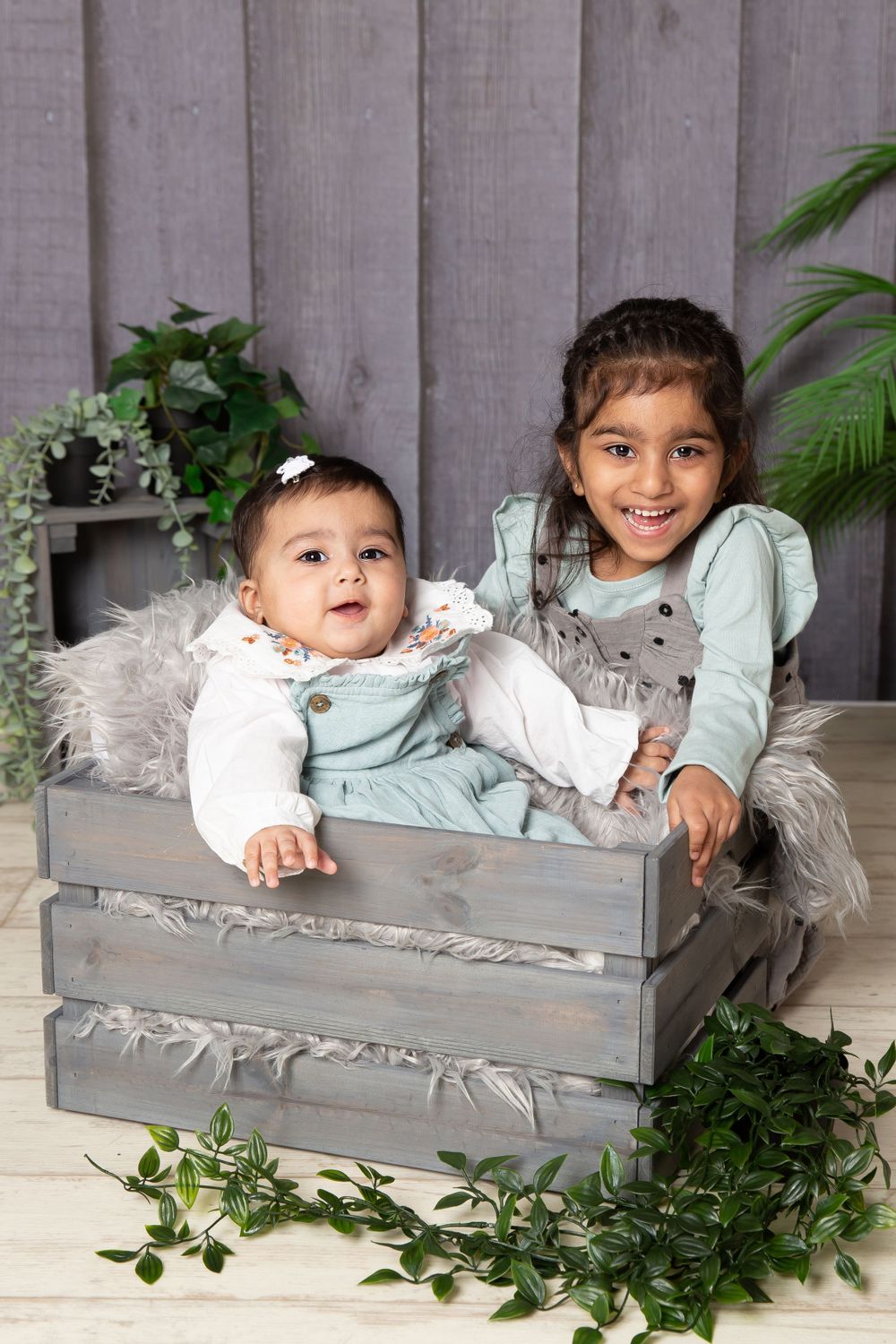 2 little girls in green with baby in a grey crate on a grey wooden background