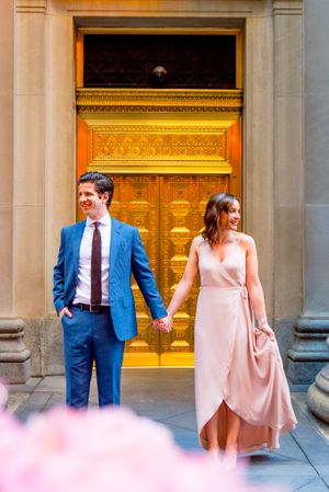 man in navy suit and woman in pink dress hold hands and look out to the sides in front of gold door in downtown Chicago