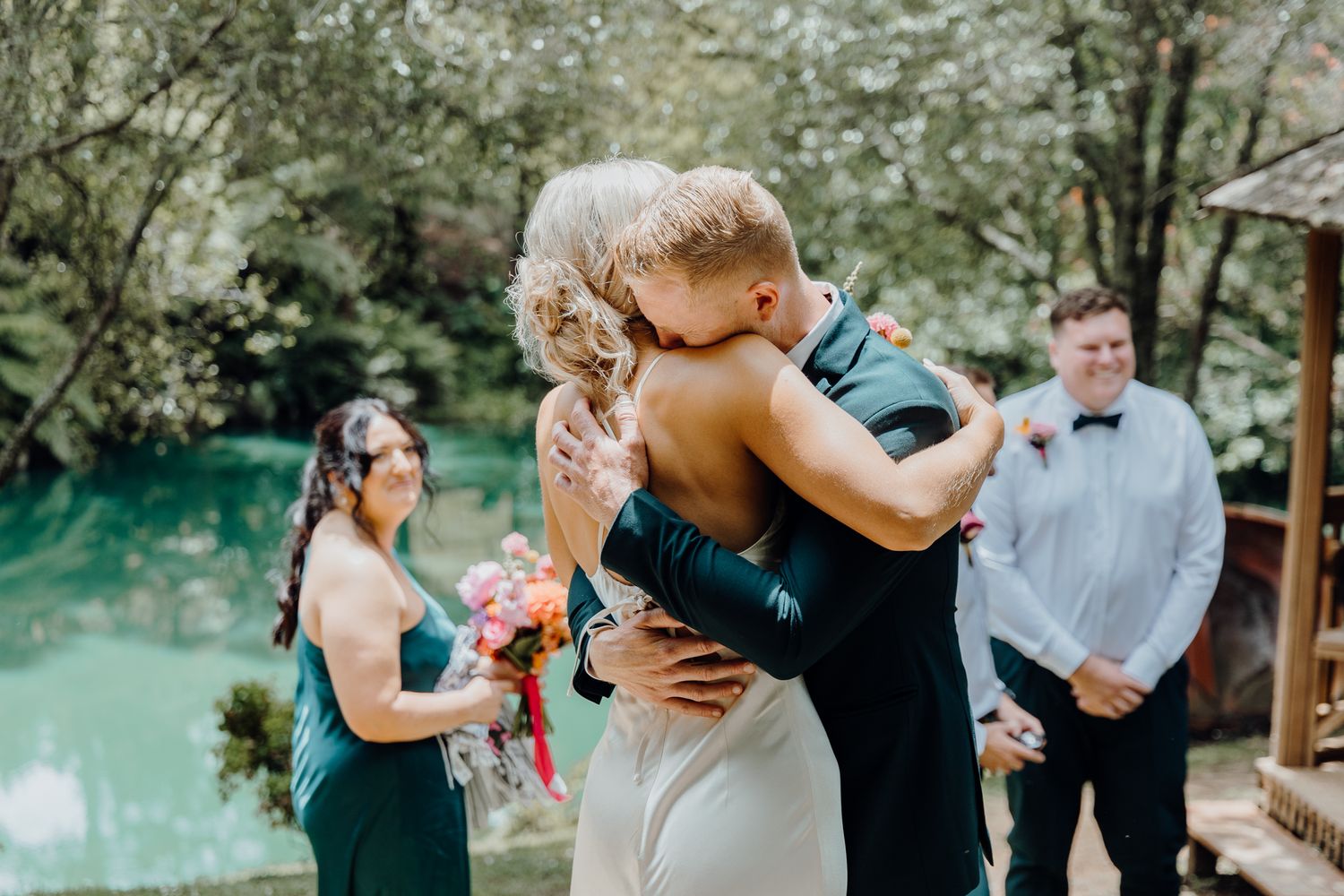 Wedding couple embraces during an outdoor ceremony by a tranquil pond with attendants and guests looking on.