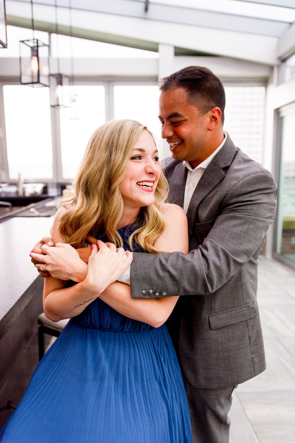 young woman with blonde hair smiles as man in grey suit hugs her from behind on the rooftop of Lytle Park Hotel