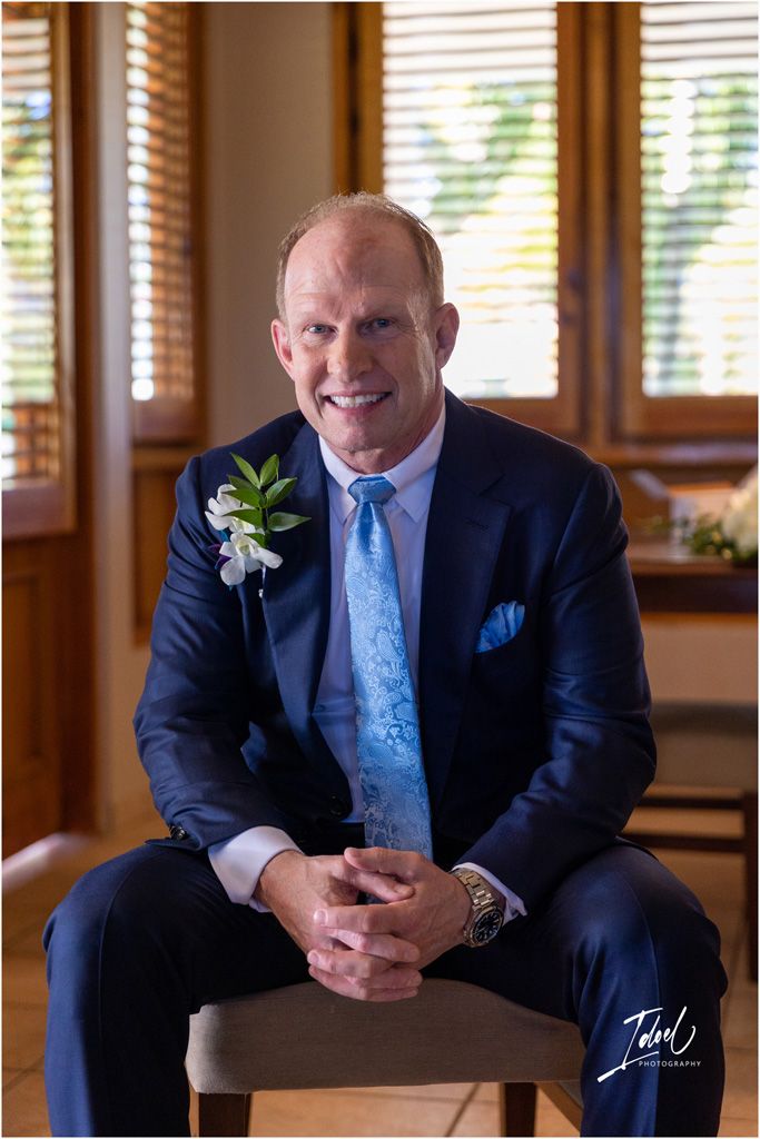 A suited figure wearing a boutonniere smiles while seated in an elegant room with wooden windows.