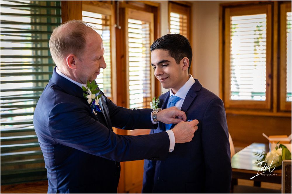 Two people in formal attire share a moment while adjusting boutonnieres before a ceremony.
