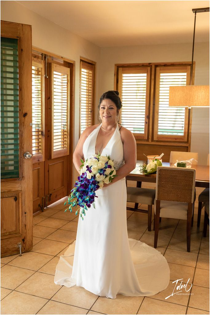 A bride in an elegant white dress poses with her blue and white floral bouquet near wooden windows.