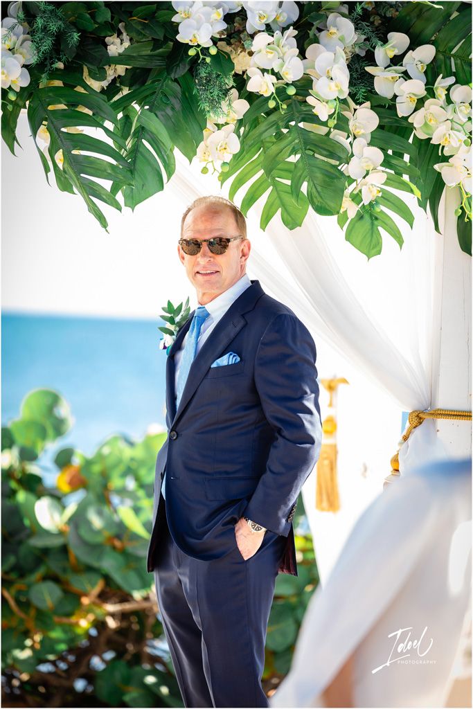 A suited figure poses under tropical foliage and white orchid arch with ocean view.