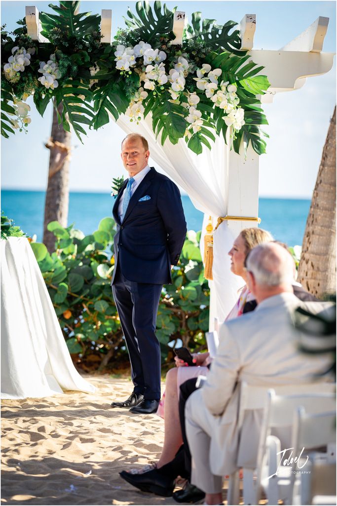 A beachside wedding ceremony setup with tropical monstera leaves and white floral arch against the ocean backdrop.