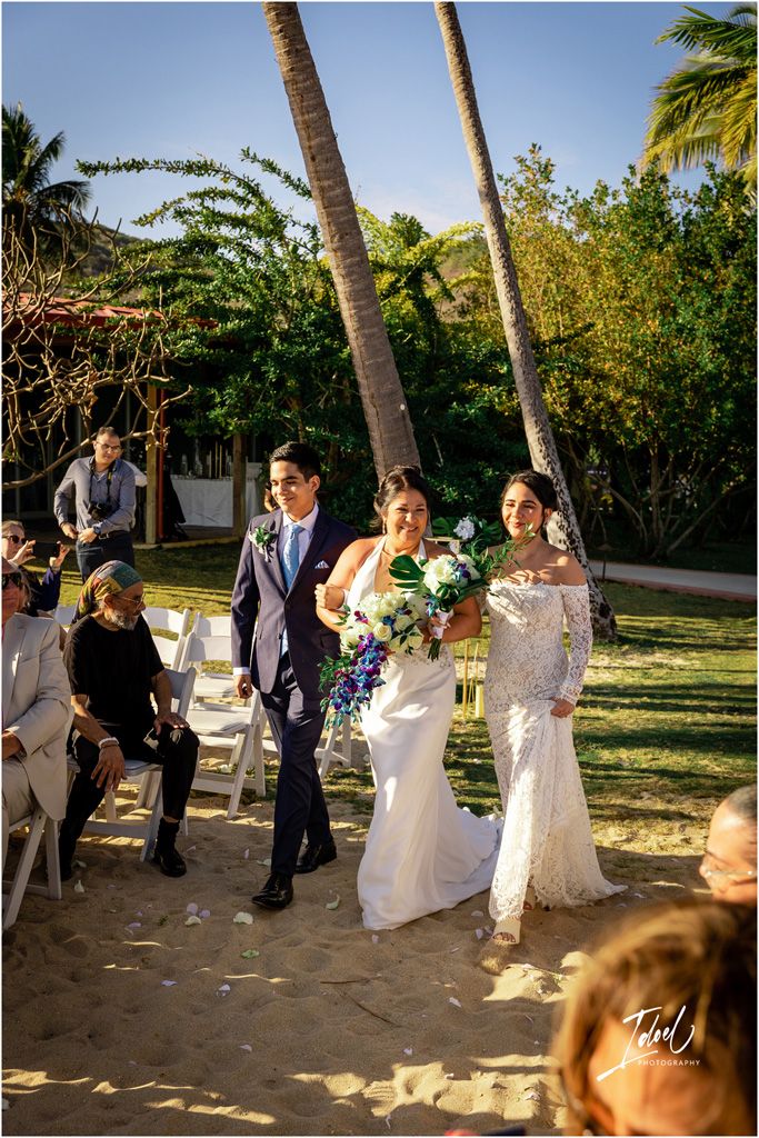 Wedding party walking down a palm-lined path during an outdoor garden ceremony.