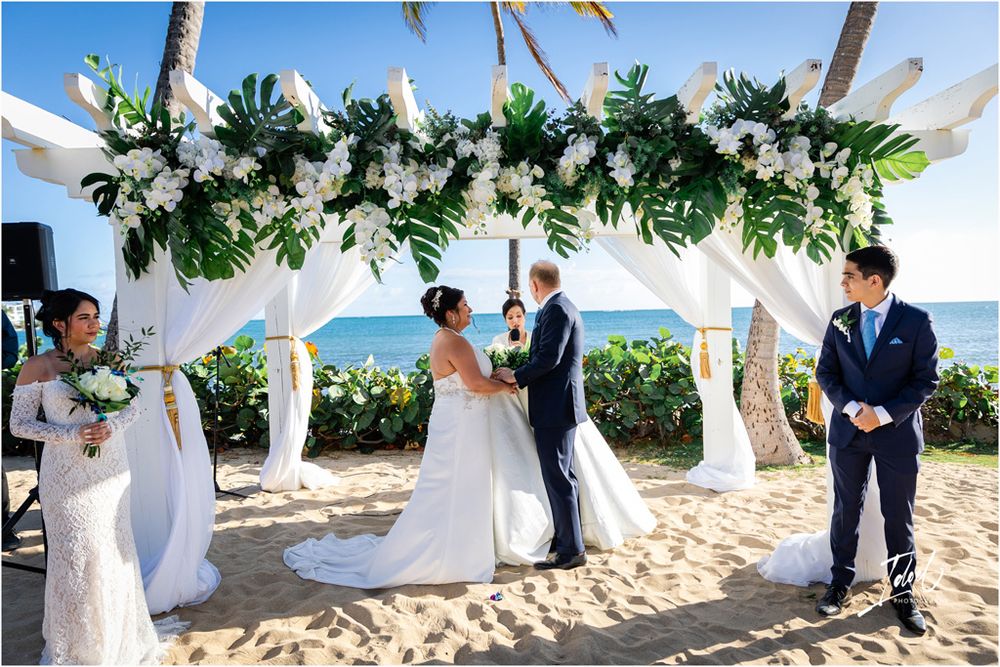 Tropical beach wedding ceremony with monstera leaf and white flower arch overlooking turquoise ocean waters.