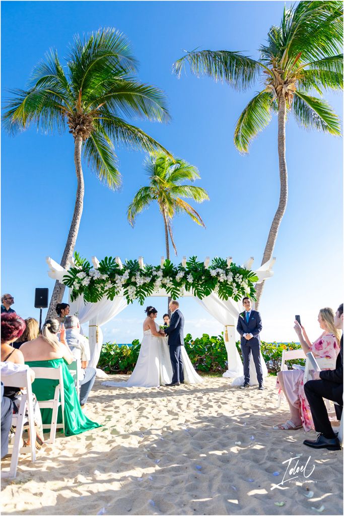 Beach wedding ceremony under palm trees with guests seated on white chairs watching the exchange of vows.