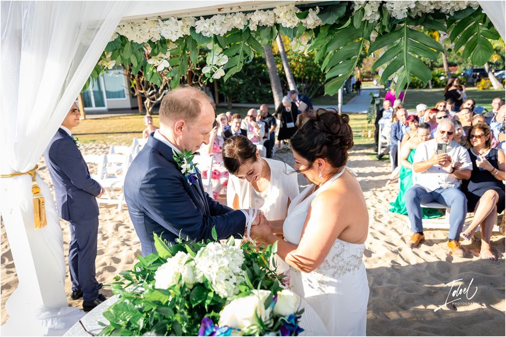 A wedding ceremony on sandy beach with tropical floral decorations and white draped fabric.