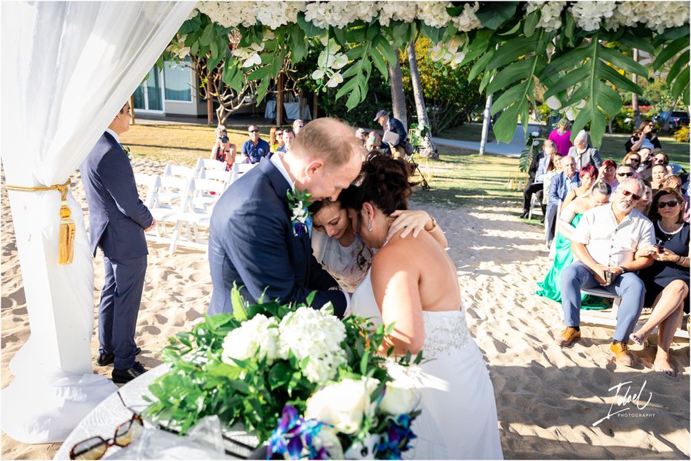 An emotional exchange during a beach wedding ceremony under a decorated floral arch.