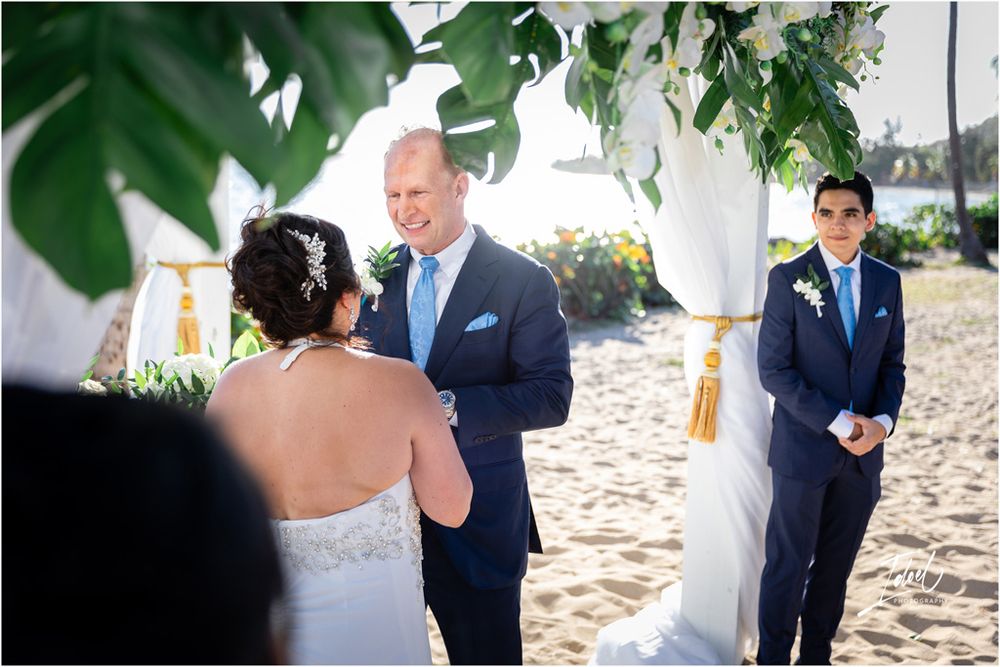 Beachside wedding ceremony with tropical greenery arch and attendants in navy suits.