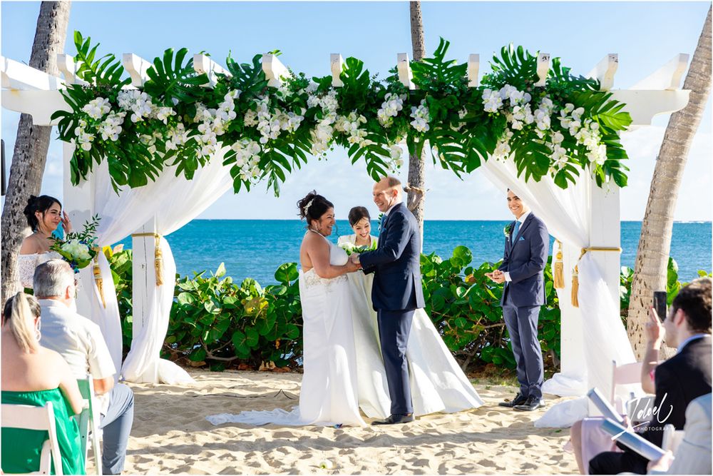 Wedding ceremony on beach with monstera leaf arch, white draping, and turquoise ocean views.