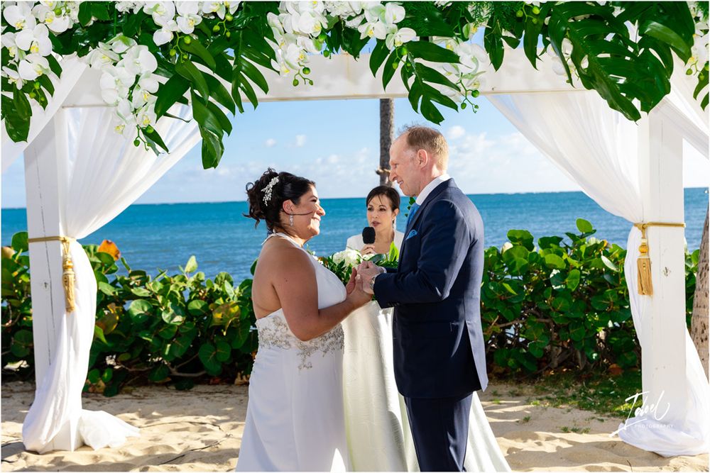 A beachside wedding ceremony under a white draped arch with tropical greenery and flowers overlooking the ocean.